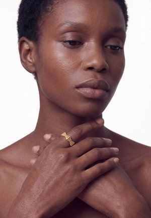 Mujer con cabello corto tocándose suavemente el cuello y el pecho, lleva un anillo dorado texturizado en el dedo anular de la mano derecha.