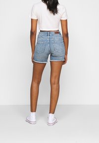 Žena stojící zády, oblečená v světlých džínových krátkých kalhotách s vysokým pasem, bílém crop-top tričku a bílých vysokých teniskách Converse.