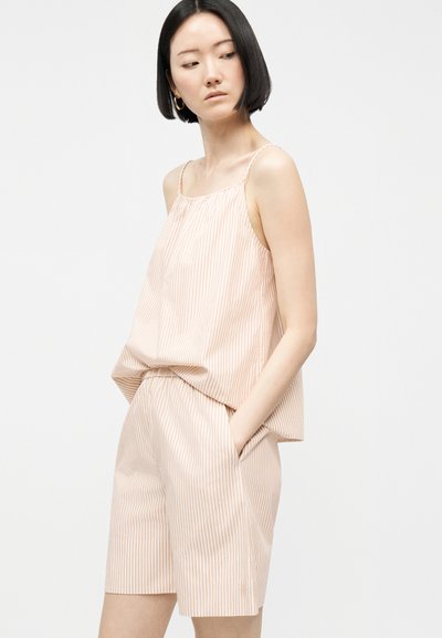 Žena s krátkymi čiernymi vlasmi, oblečená v béžovom a bielo pruhovanom bezrukávovom topu a zodpovedajúcich krátkych nohaviciach, stojaca s rukami v vreckách.