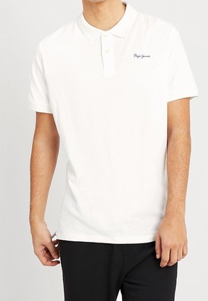 Mężczyzna w białej, krótkorękawowej koszulce polo z małym granatowym logo na lewej stronie klatki piersiowej, w połączeniu z czarnymi spodniami.