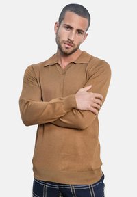 Homme aux cheveux courts et foncés et à la barbe, portant un pull marron à manches longues et un pantalon bleu à carreaux, les bras croisés, regardant la caméra.