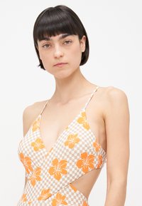 Eénstuk zwempak met een diepecolleté, met een oranje bloemenpatroon op een geblokte beige achtergrond. Dunne bandjes en uitsnijding.
