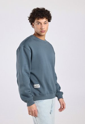 Jonge man met krullend haar, gekleed in een blauwgrijze trui en lichtblauwe spijkerbroek, staand tegen een effen lichte achtergrond.