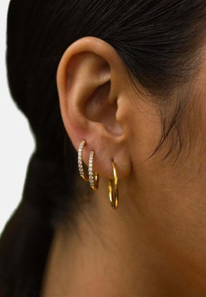 Nahaufnahme eines linken Ohrs mit drei goldenen Creolen, zwei davon mit kleinen eingearbeiteten Diamanten und eine schlichte, vor einem dunklen Haarhintergrund.