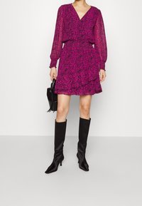 Vestido de manga larga con escote en V profundo en fucsia con un patrón abstracto negro, cintura ceñida, falda con volantes, combinado con botas negras altas hasta la rodilla.