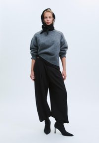 Maglione grigio in lana con scollo rotondo, pantaloni drappeggiati neri e stivaletti neri con tacco. Il modello indossa una sciarpa nera annodata intorno al collo.