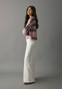 Suéter de punto a rayas rosas y grises combinado con pantalones acampanados blancos que tienen tachuelas en los lados. La modelo está de lado frente a un fondo gris.