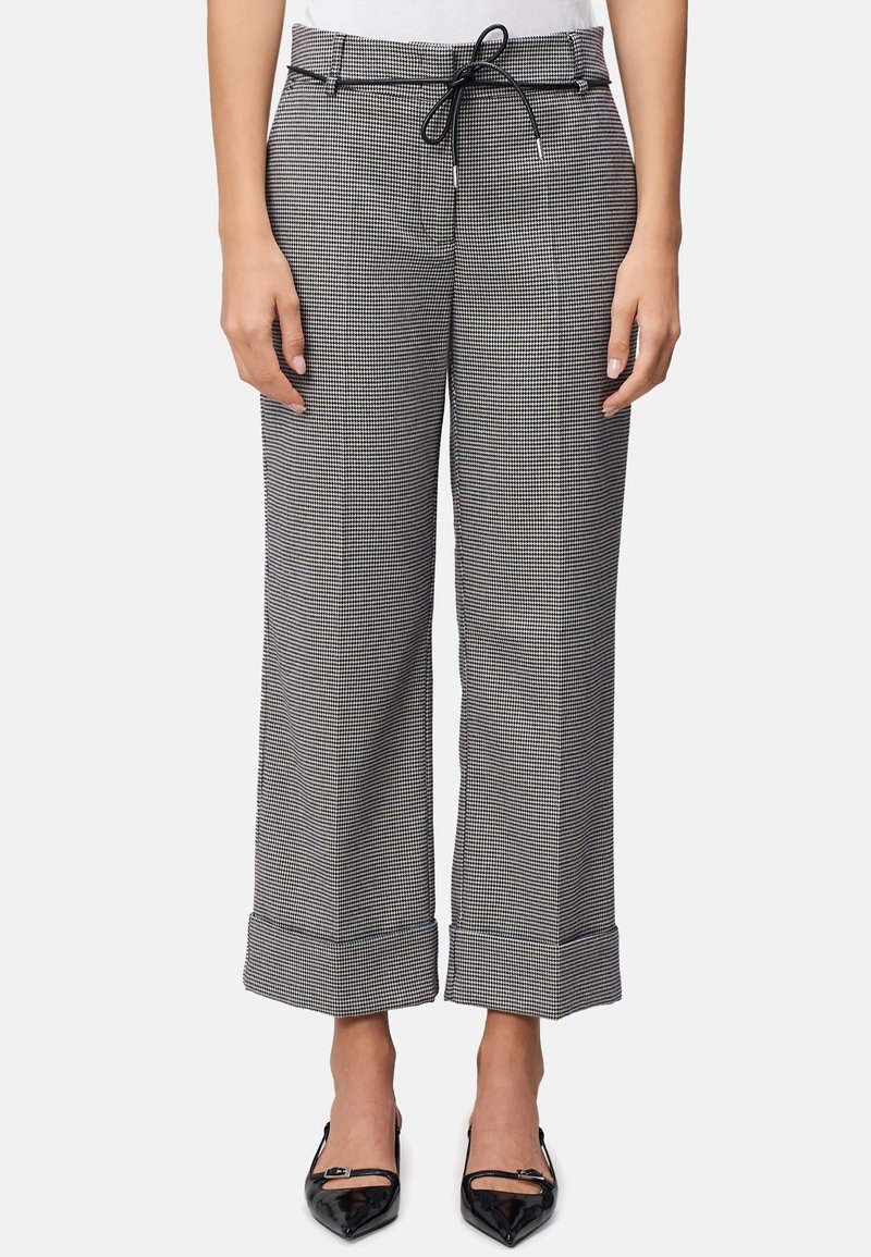 Femme portant un pantalon large court à carreaux noirs et blancs avec des ourlets retournés et une fine ceinture noire, associé à des chaussures noires à bouts pointus.