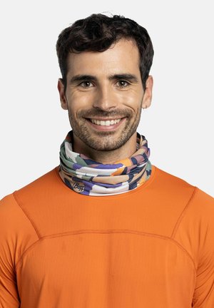 Hombre sonriente con cabello oscuro que lleva un pañuelo de cuello con patrón colorido y una camisa naranja de manga larga contra un fondo claro y liso.