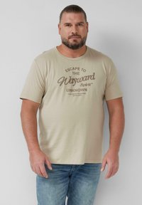 Beige katoenen t-shirt met korte mouwen, voorzien van een bruine grafische print met de tekst "Ontsnap naar de Onbekende Dwaalmolen." Draagt met lichtblauwe jeans.