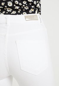 Jeans in denim bianco con una tasca posteriore e un'etichetta metallica. La texture è liscia e il design presenta una vestibilità classica e dettagli di cucitura sottili.