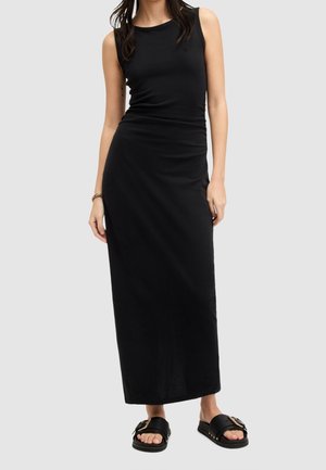 Kobieta w czarnej, bezrękawnikowej sukni maxi i czarnych sandałach na płaskiej podeszwie z złotą klamrą, stojąca na tle jednolitego, jasnoszarego tła.