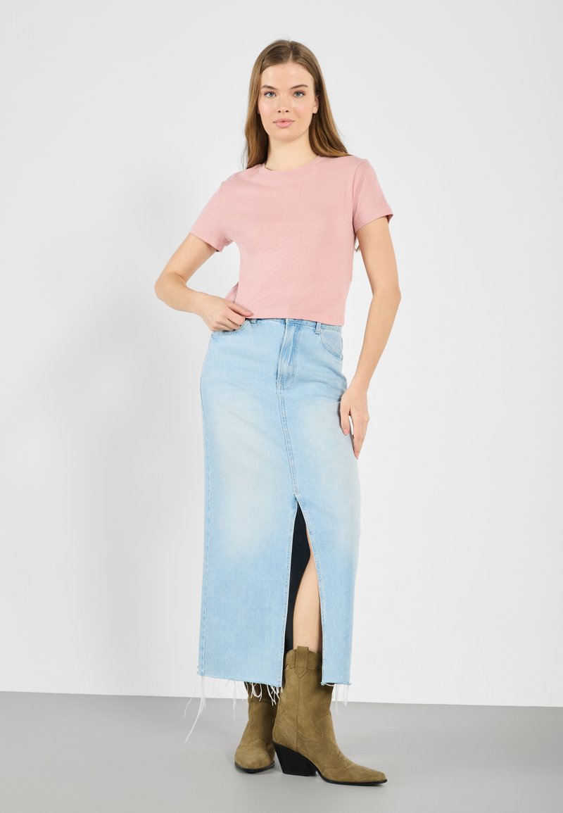 Lichtroze shirt met korte mouwen, gecombineerd met een lichtblauwe denim maxi rok met hoge taille, voorzien van een split aan de voorkant en een rafelige zoom. Bruine haklaarzen.