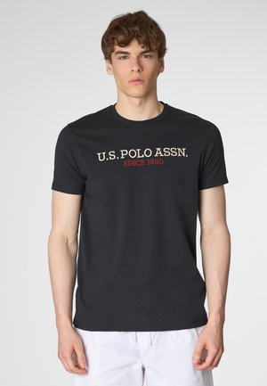 Un giovane con i capelli ricci indossa una maglietta scura della U.S. Polo Assn. Lo sfondo è grigio, concentrando l'attenzione sulla sua espressione e sul suo abbigliamento informale.