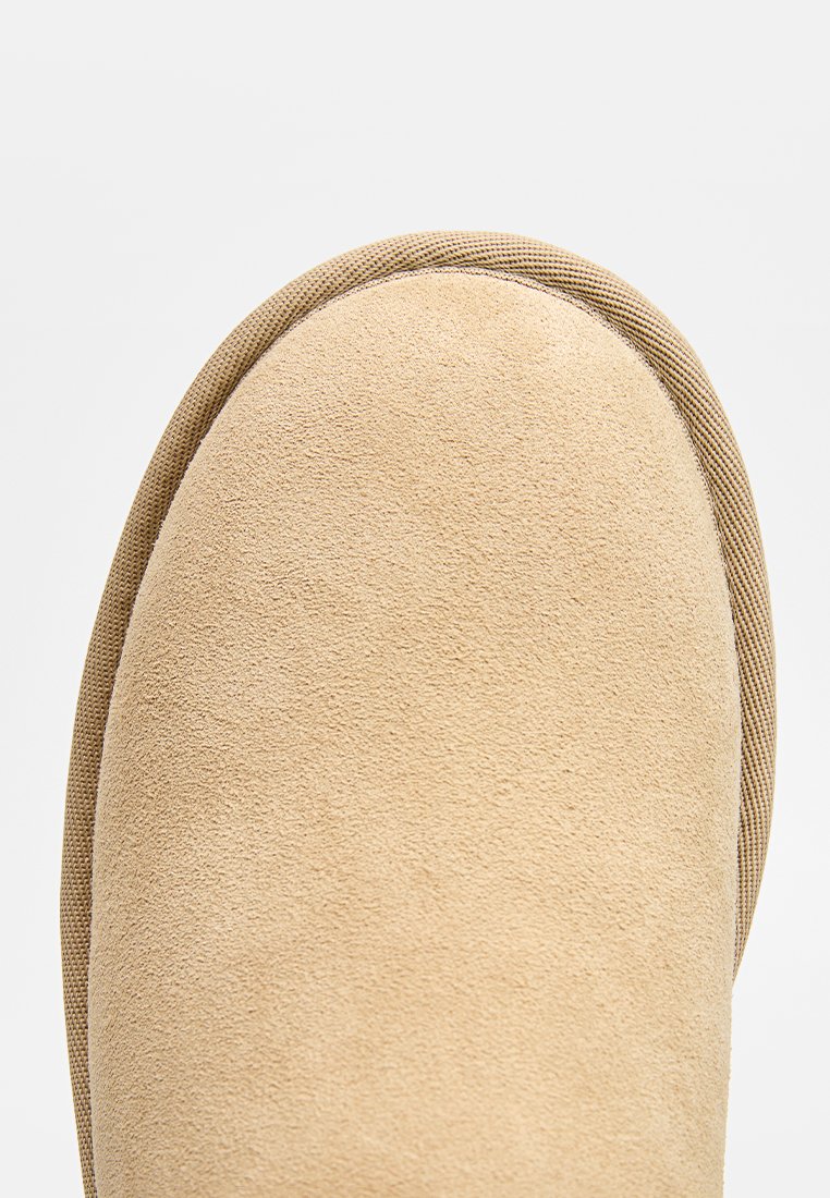 Beige mockakänga med rund tå, texturerad yta och kontrasterande tan gummisole. Sidodetaljerna inkluderar subtil sömnad.
