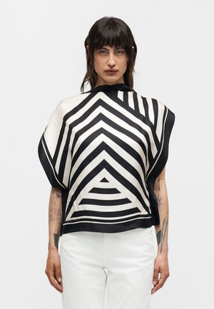 Femme portant un haut à motifs géométriques noir et blanc avec des rayures triangulaires, associé à un pantalon blanc, debout sur un fond uni.