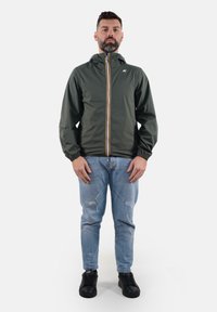 Giacca zip-up verde scuro con cappuccio, polsini elasticizzati e zip gialla a contrasto. Indossata su jeans chiari strappati e sneaker nere.