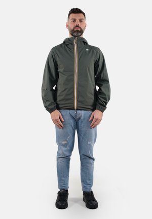 Giacca zip-up verde scuro con cappuccio, polsini elasticizzati e zip gialla a contrasto. Indossata su jeans chiari strappati e sneaker nere.