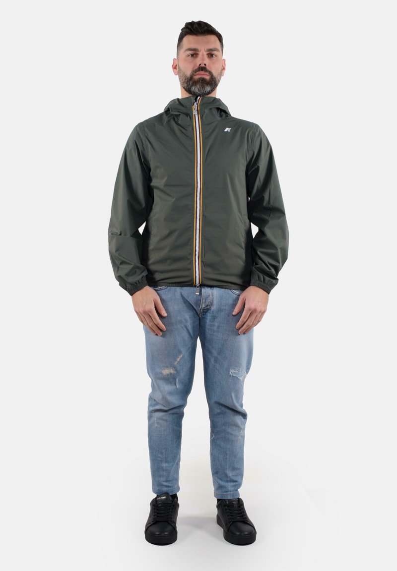 Giacca zip-up verde scuro con cappuccio, polsini elasticizzati e zip gialla a contrasto. Indossata su jeans chiari strappati e sneaker nere.