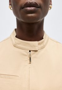 Άτομο με σκουρόχρωμο δέρμα που φοράει χρυσούς κρίκους στα αυτιά και μπεζ τοπ με φερμουάρ και όρθιο γιακά, με κλειστά χείλη, μπροστά σε απλό φόντο.