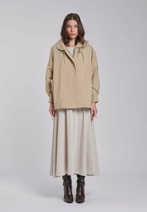 Kobieta stojąca, ubrana w beżową, luźną kurtkę, długą kremową spódnicę i ciemnobrązowe kozaki sięgające kolan na jednolitym jasnym tle.