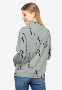 Szara bluzka z długim rękawem, z dużymi czarnymi i zielonymi literami w różnych czcionkach. Tkanina wydaje się miękka, z luźnym krojem i bocznymi rozcięciami.