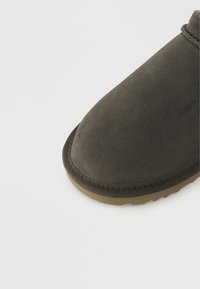 Σκούρο γκρι παντόφλα από σουέτ με στρογγυλεμένη μύτη, ταμπά λαστιχένια σόλα και ορατά ράμματα κατά μήκος των άκρων. Ομαλή υφή χωρίς διακοσμήσεις.