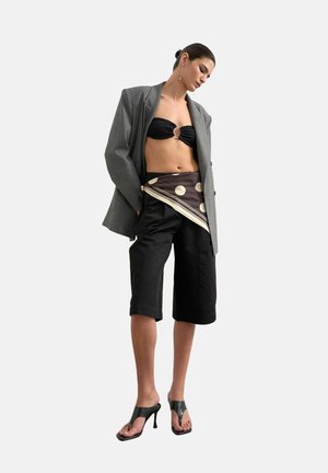 Vrouw draagt een oversized grijze blazer, een zwarte bandeau top, een polkadot sjaal over zwarte knielange shorts en zwarte sandalen met hakken.