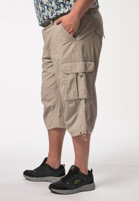 Shorts cargo beiges avec plusieurs poches, ourlets à cordon de serrage, fabriqués à partir d'un tissu léger. Associés à des chaussures de sport noires avec des accents jaunes.
