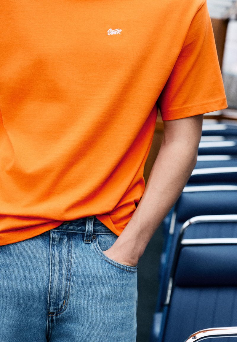 Personne portant une chemise orange à manches courtes et un jean bleu, une main dans la poche, debout à côté de chaises bleues et argentées.