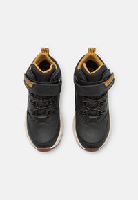 Černé a hnědé dětské tenisky s bílými podešvemi, suchým zipem a tkaničkami, zobrazené shora na bílém pozadí.