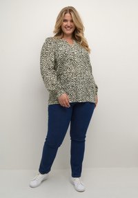 Leende kvinna står i en blus med beige och svart mönster, mörkblå jeans och vita sneakers mot en enkel bakgrund.