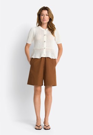 Femeie îmbrăcată într-un top tricotat alb cu mâneci scurte și nasturi și pantaloni scurți maro până la genunchi, stând cu mâinile în buzunare pe un fundal alb.