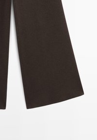 Pantalons larges à jambes marron foncé avec ourlets nets sur fond blanc, montrant la texture du tissu et la coupe droite.