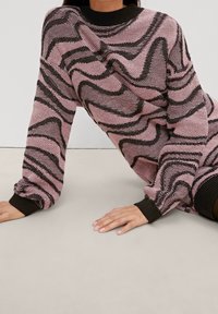 Pull en tricot avec une base rose et des motifs ondulés noirs, à manches longues, avec un col et des poignets côtelés noirs. Tissu texturé.