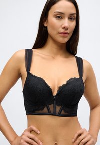 Czarny koronkowy biustonosz z przezroczystymi ramiączkami, głębokim dekoltem i falistymi krawędziami. Posiada zapięcie z przodu oraz ozdobne siateczkowe detale na pasku.