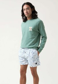 Sudadera verde con la palabra "SURF" estampada, combinada con pantalones cortos blancos que presentan patrones gráficos en azul de sol, olas y plantas. Diseño casual.