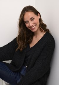 Schwarze, gestrickte Strickjacke, die über einem dunklen Hemd getragen wird, mit langen Ärmeln und offenem Vorderteil. Der Stoff scheint fuzzy und strukturiert zu sein.