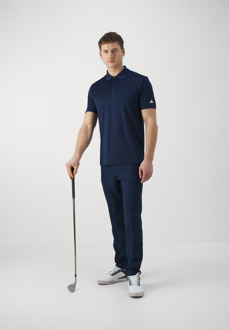 Polo shirt bleu marine avec patte de boutonnage à trois boutons et logo sur la manche, associé à un pantalon bleu marine et des chaussures de golf blanches. Club de golf tenu à la main.