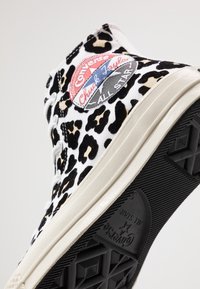 Zapatillas Chuck Taylor de Converse de caña alta en una tela blanca con estampado de leopardo negro y marrón. Presenta un parche de logo en rojo y azul y suela de goma.