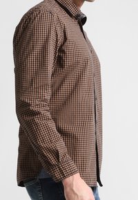 Camisa de manga larga a cuadros marrón y negro con cuello abotonado, diseño ajustado y textura suave de algodón.