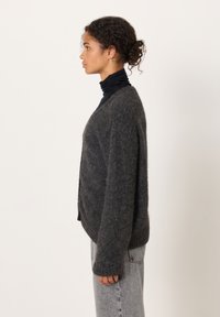 Dark gray cardigan with a loose fit, knit texture, and long sleeves, paired with a black turtleneck and light gray jeans.