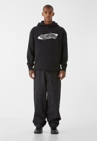 Sudadera negra con el logotipo "Starboy" en blanco, bolsillo frontal y puños acanalados. Combinada con pantalones de mezclilla oscuros y holgados y zapatos negros.