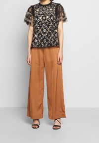 Sort blomstret broderet top med gennemsigtige puffede ærmer, parret med brune bredbenede bukser og sorte strophæle.