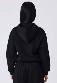 Personne avec de longs cheveux noirs, de dos, portant un sweat à capuche noir et un pantalon noir assorti sur un fond clair.