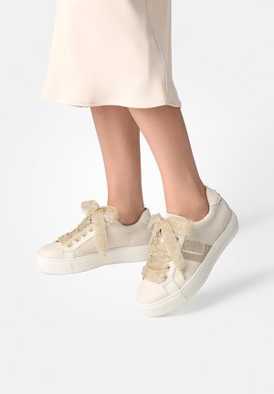 Beige platform sneakers with glittery gold laces worn with a light cream skirt, showing ankles and lower legs against a white background.