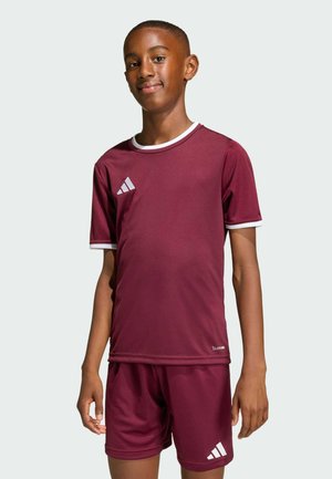 Chłopiec stojący, ubrany w bordowy sportowy strój Adidas z białym wykończeniem, lekko uśmiechający się na jednolitym jasnym tle.