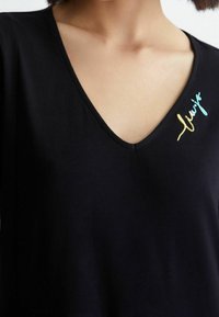 Mustat V-päällyskäytävät T-paita lyhyillä hihoilla, jossa on värikkäitä brodeerattuja tekstejä vasemmalla noin kaula-aukon kohdalla. Pehmeä, sileä kankaankudoksen rakenne.