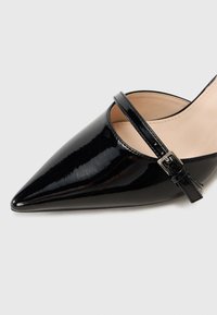 Scarpa nera in vernice con tacco alto, punta affusolata e cinturino alla caviglia con fibbia argentata. Texture liscia e design elegante.