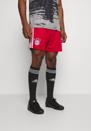 Mężczyzna w czerwonych szortach piłkarskich FC Bayern Monachium, czarnych podkolanówkach Adidas i czarnych korkach piłkarskich, stojący na białym tle.